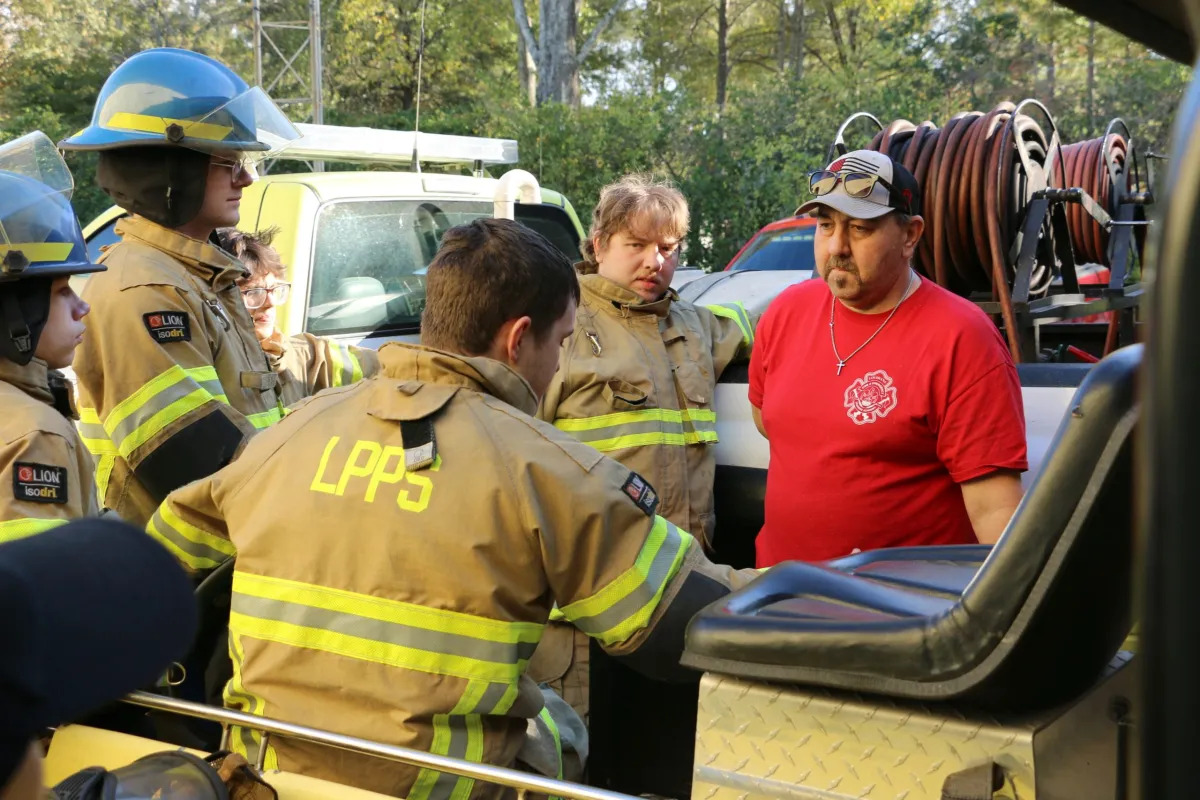 High school career and tech education: Five men and students wearing tan firefighting gear with yellow reflective strips sitting in a fire truck with rolls of fire hoses in the background