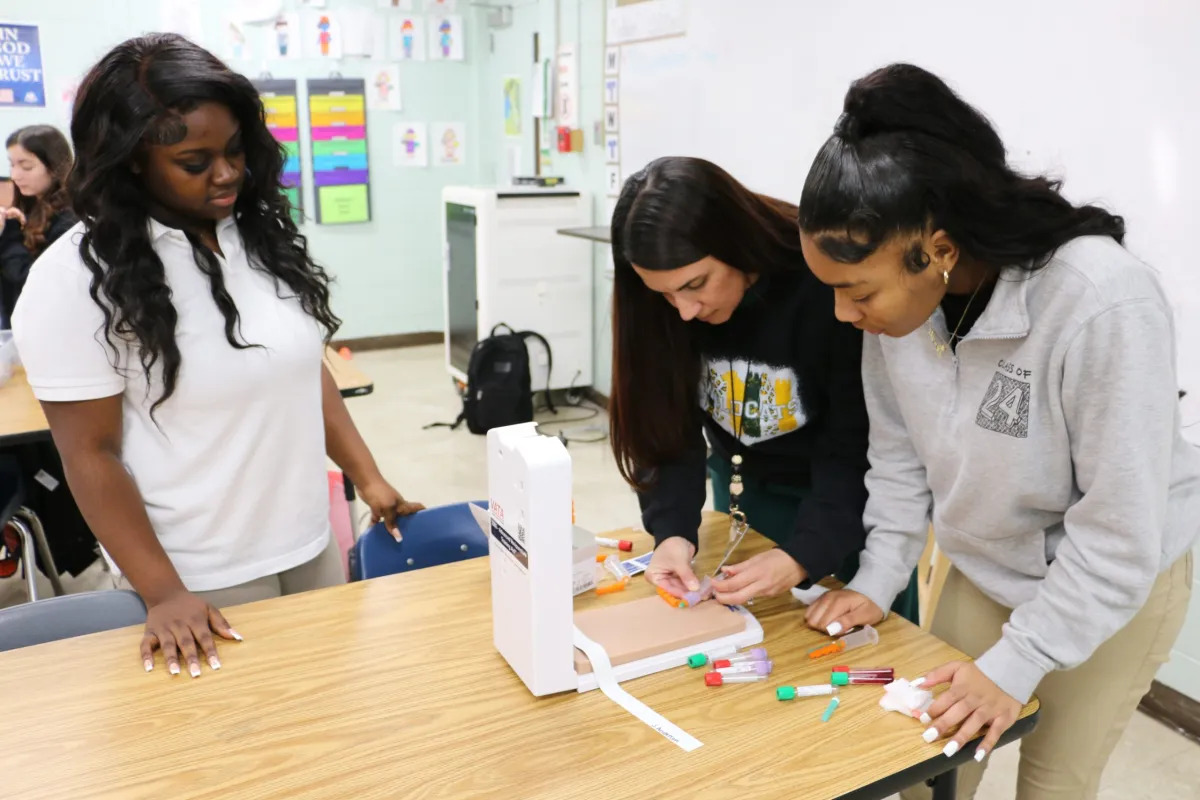 High school career and tech education: Three female high school students with long, black hair in casual clothing stand sorting vails with various colored tops on a table in a classroom