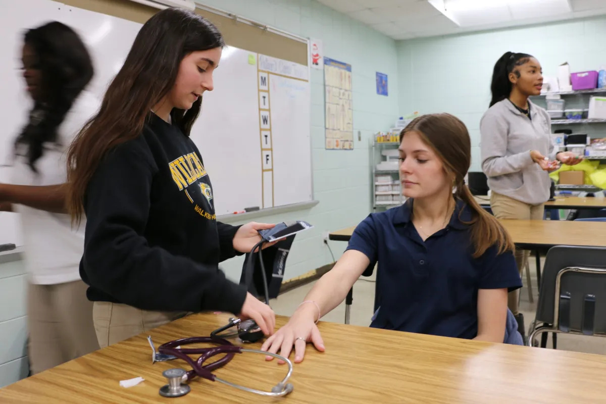 High school career and tech education: Female student with long dark hair stands at table with a stethoscope while another female sits at the table and two other students stand in the background