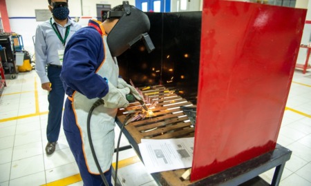 Person with welding equipment and protective eye/headgear works in a welding booth inside a classroom, with a male teacher observing in background