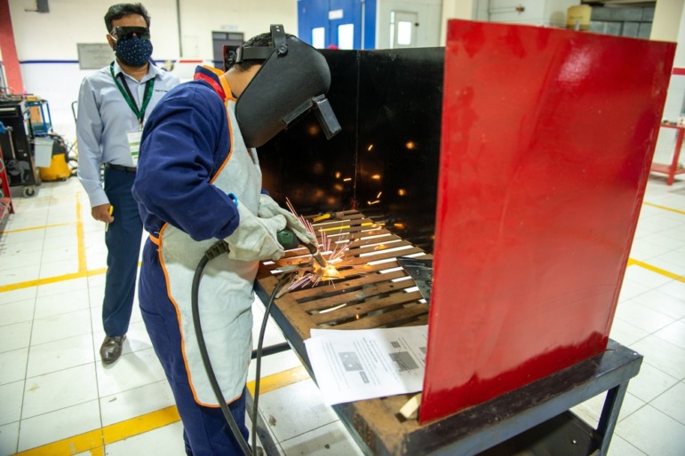 education, Person with welding equipment and protective eye/headgear works in a welding booth inside a classroom, with a male teacher observing in background