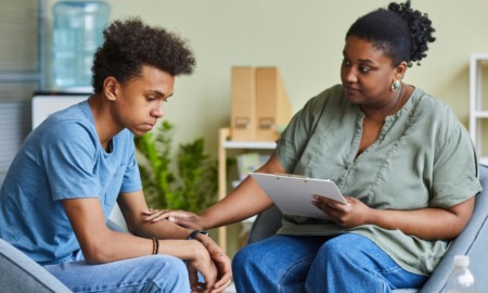 Teen Therapy: Black woman with cark hair pulled back in gray shirt and bluejeans sits holding hand of seated Black teen boy with black hair in blue shirt and bluejeans who looks despondent