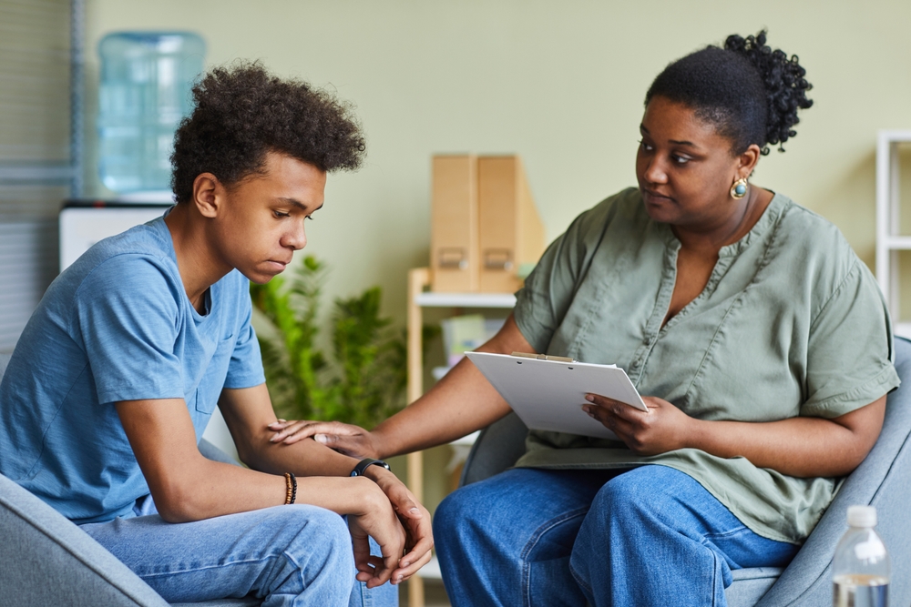 Teen Therapy: Black woman with cark hair pulled back in gray shirt and bluejeans sits holding hand of seated Black teen boy with black hair in blue shirt and bluejeans who looks despondent