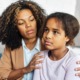 social and emotional learning, SEL: Black teen girl with brown hair in white top sits next to Black woman with brown hair in tan jacket and white top on light couch. Woman holds girl around shoulders.