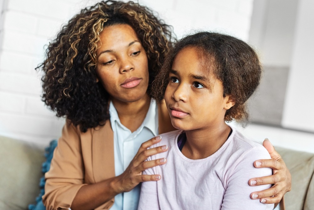 social and emotional learning, SEL: Black teen girl with brown hair in white top sits next to Black woman with brown hair in tan jacket and white top on light couch. Woman holds girl around shoulders.