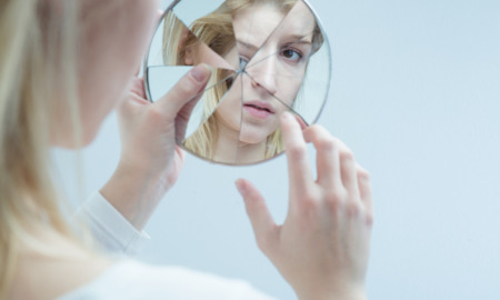 Teen mental health: Young blonde woman looking at her reflection in a broken mirror