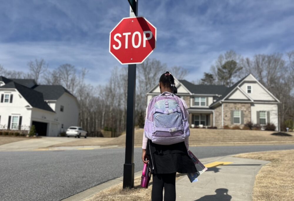 Georgia School Voucher Bill: Young Black girl with light lavender and blue backpack, wearing black pants stands on sidewalk next to red stop sign with back to camera and large houses in the background.
