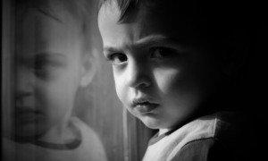 Child Care Research: Closeup in black and white of young boy next to window scowling into camera with face reflected in a window