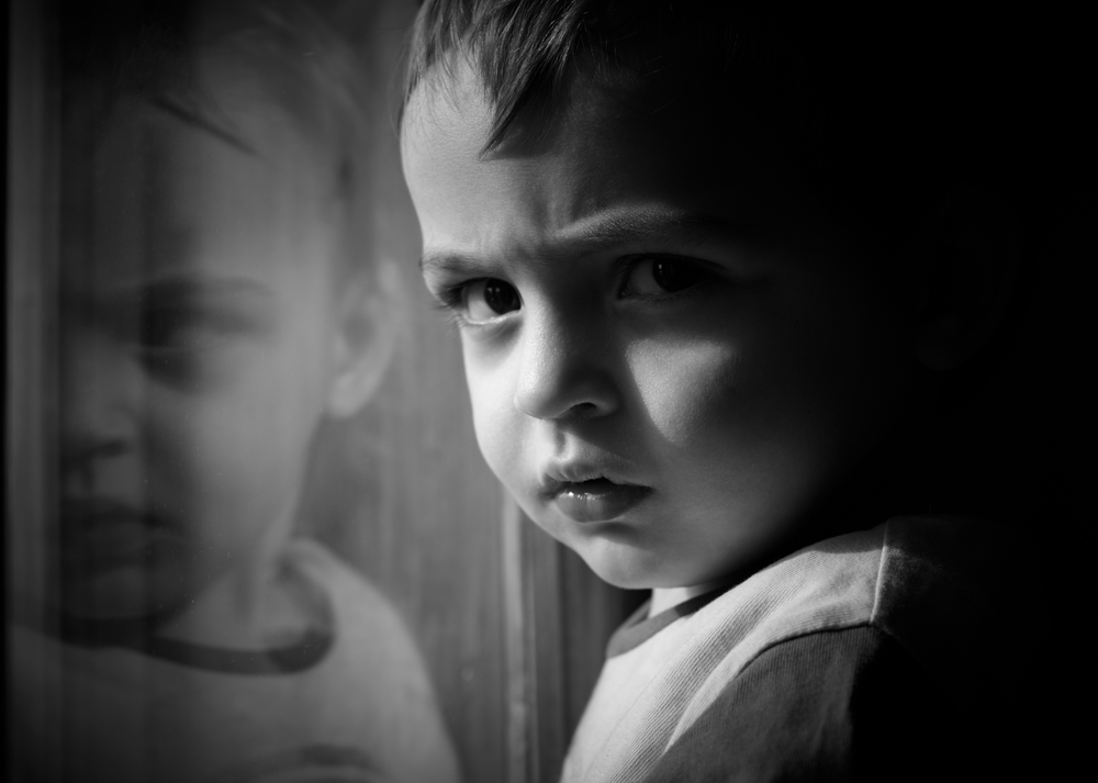 Child Care Research: Closeup in black and white of young boy next to window scowling into camera with face reflected in a window