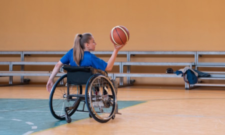 Adaptive sports: Teen with long blonde ponytail sitting in wheelchair on indoor basketball court holds basketball up in air in right hand