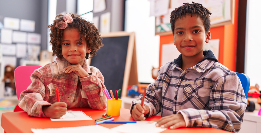 Black charter school: Two young Black children in plaid shirts sit next to each other at a school desk drawing on paper smiling into camera