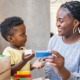Black women are still holding up the child care industry: Black woman with long hair plays blocks with young black boy in child care setting