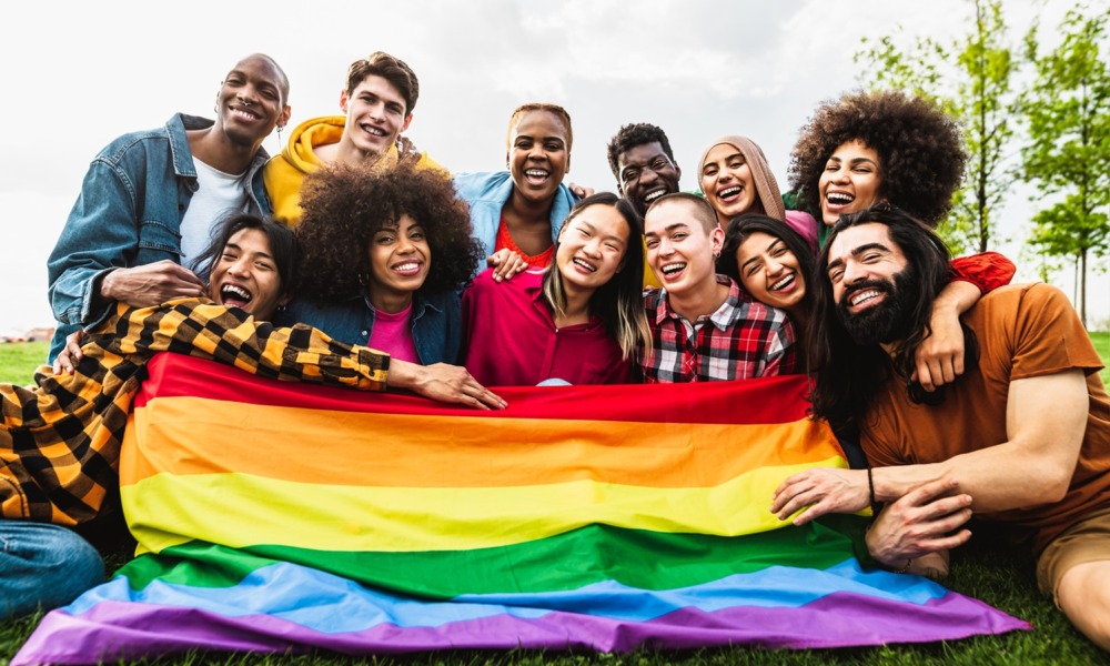 supporting queer youth, LGBTQ+ community: group of smiling, diverse youth with rainbow flag draped in front of them