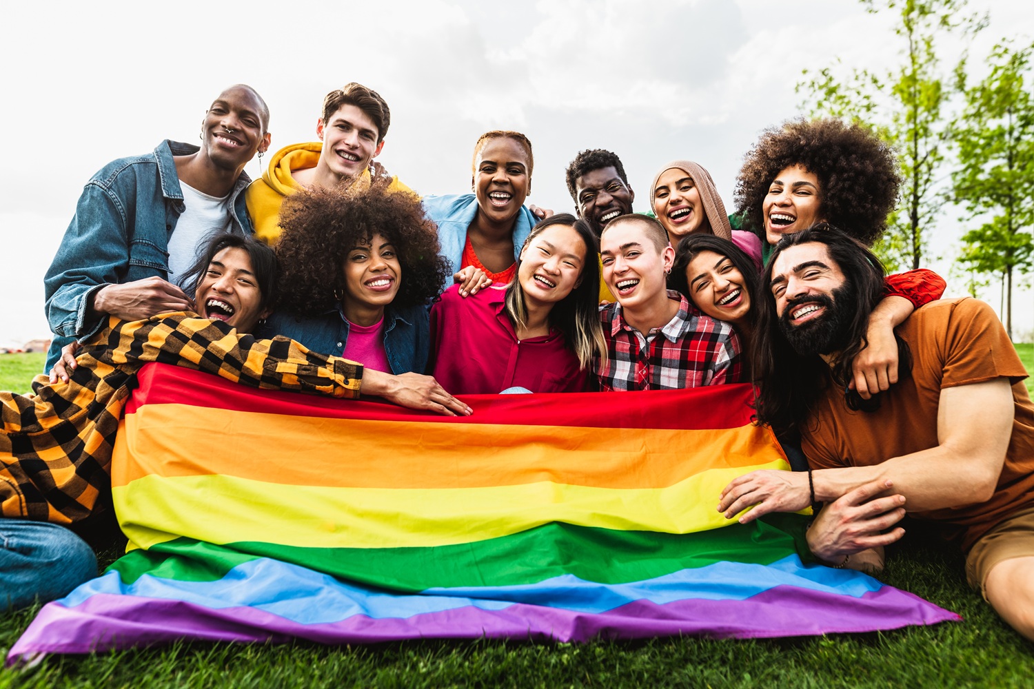 supporting queer youth, LGBTQ+ community: group of smiling, diverse youth with rainbow flag draped in front of them