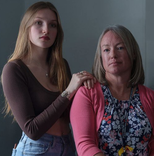 Student-led mental health: Long-haired, blonde teen girl in brown shirt and jeans, stands with hands on shoulders of seated, middle aged woman with gray hair in bright pink sweater
