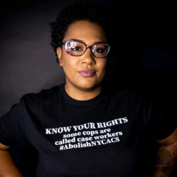 Lawsuit NYC ACS Investigation: Black woman wirh shirt natural black hair and large, black-framed glasses wearing black t-shirt with white text stares stoically into camera