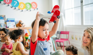 Child-Care-Guidelines: Toddler boy with short dark hair in denim overalls and red shirt holds red toy airplane above his head in playroom filled with other children and two teachers sitting on the floor