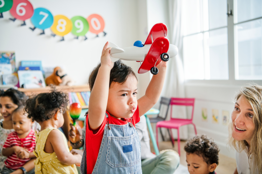 Child-Care-Guidelines: Toddler boy with short dark hair in denim overalls and red shirt holds red toy airplane above his head in playroom filled with other children and two teachers sitting on the floor