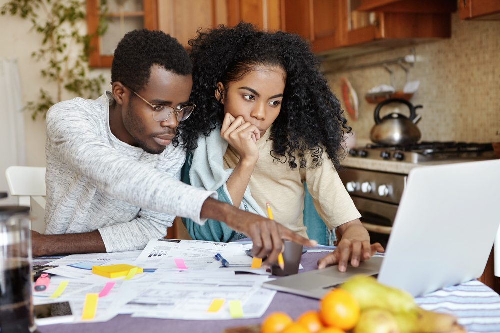 College loans: Black man and young woman sit at kitchen table covered with forms looking at laptop screen. Man id pointing at screen. Kitchen counter and cupboards are in background