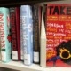 Georgia targets ALA: A row of multicolored books are lined up on a plain shelf. All have their spines facing the camera. On the right, the book "Take the Mic: Fictional Stories of Everyday Resistance" is turned so the cover faces the camera. The cover is red, the title is in black and white, and it features an illustration of a side profile of a Black woman yelling into a microphone that is on fire. There are 11 authors whose names are scattered across the cover.