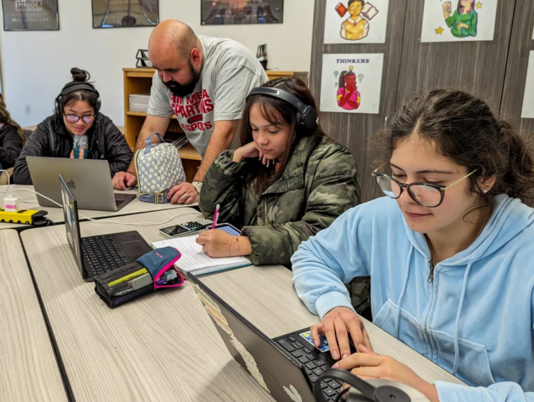 Data Science Math: Three high school students sit at laptops in classroom with male teacher standing behind one assisting them.