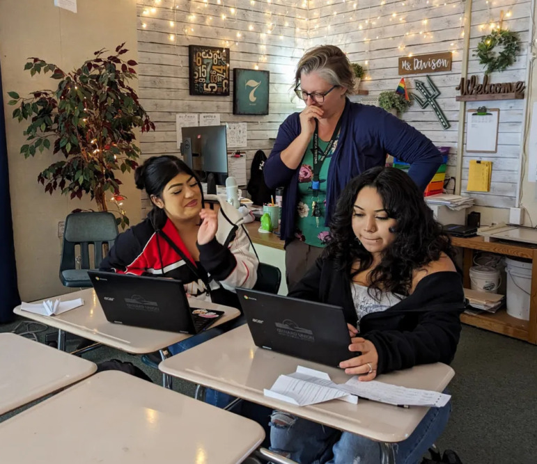 Data Science Math: Two high school students sit at laptops in classroom with female teacher standing behind them assisting them.