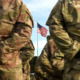 National Guard school Violence: Closeup of backs of people standing in two lines wearing camo green and brown army uniforms with an American flag in background