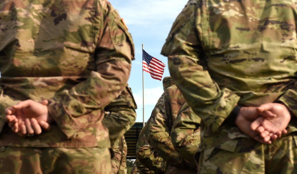 National Guard school Violence: Closeup of backs of people standing in two lines wearing camo green and brown army uniforms with an American flag in background