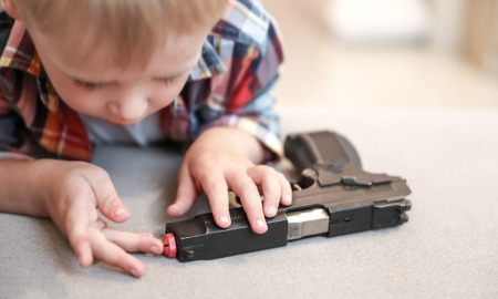 Gun Safety ar Home: Very young boy leans close to hand fun on table poking one finger into barrel.