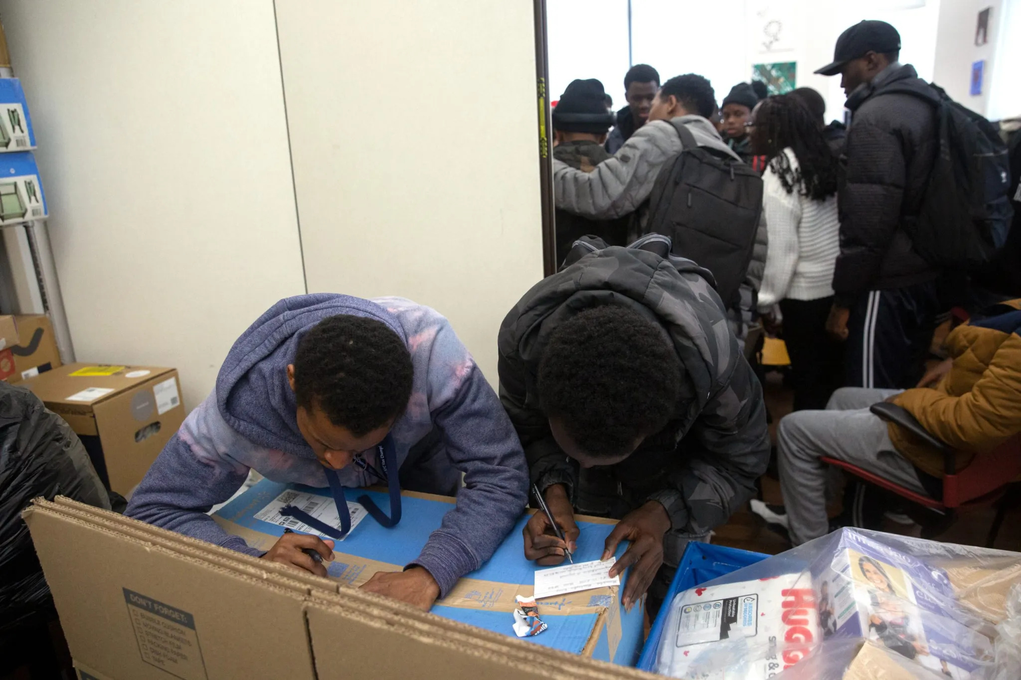 NYC homeless youth and migrants: Two black males fill out forms using the tops of large cardboard boxes for desks while several other people stand crowded together in another room in the background