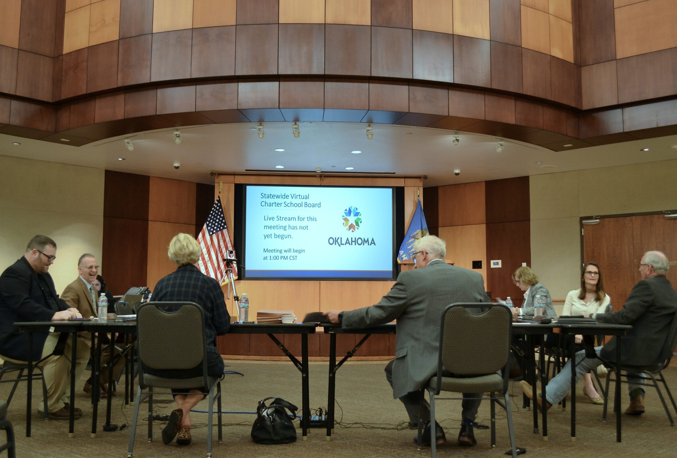 Oklahoma virtual charter schools: Several people in business-wear sit in a formal meeting room at a circle of tables, some with backs to camera, looking at a presentation screen and having a discussion .