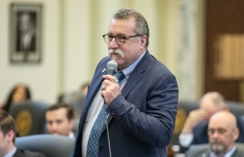 Oklahoma virtual charter schools: Middle-aged man with gray hair abd glasses in dark blue suit stands speaking into hand-held microphone in a room full of people seated around him
