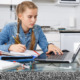 Oklahoma virtual charter schools: Teenage girl with ling blonde hair in two braids in light blue denim shirt sits in white kitchen at table with several school supplies working on laptop and writing in notebook