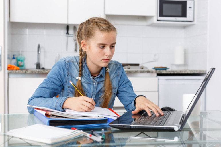 Oklahoma virtual charter schools: Teenage girl with ling blonde hair in two braids in light blue denim shirt sits in white kitchen at table with several school supplies working on laptop and writing in notebook