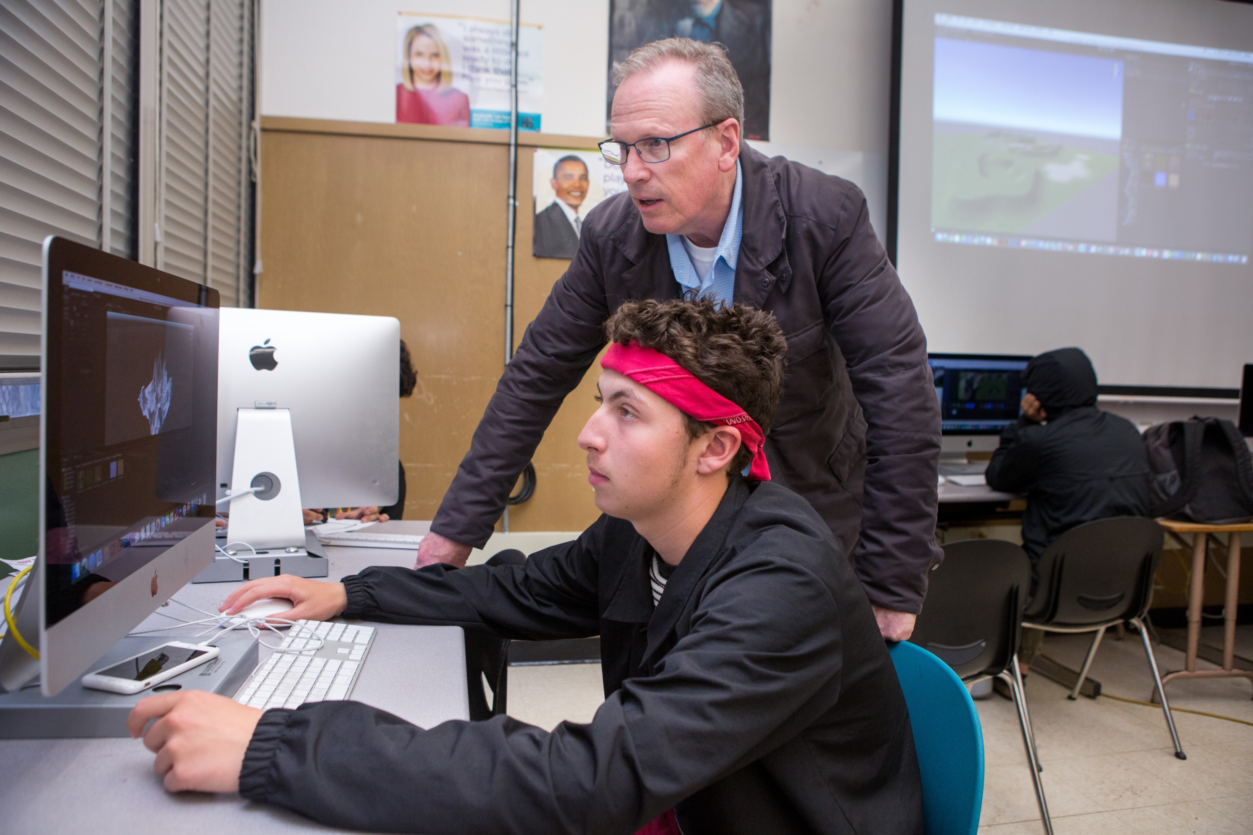Tutoring Improves Attendance: Older gray-haired man with glasses in dark suit stands beside seated teen male student with red headband and dark jacket, both looking at large monitor in computer lab