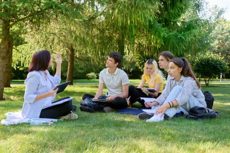 Summer learning strategies: Group of four teen students sit in circle on grass with trees in the background, all facing a teacher holding up her hand while demonstration something on laptop screen