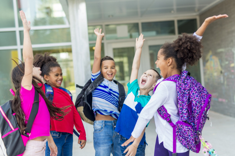 Summer learning strategies: Group of ethnically-mixed preteens with backpacks stand in circle in front of school entrance jumping with hands in air ansdsmiling at each other.