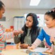 Universal prekindergarten is coming to California: woman with long, dark hair helps two young girls in preschool classroom