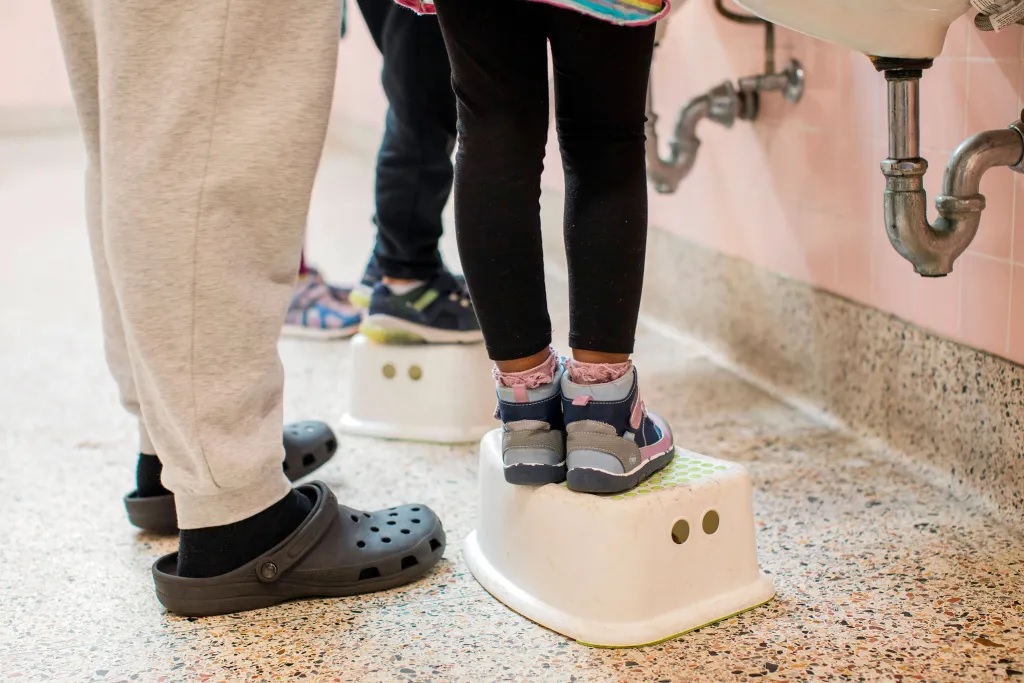 Universal prekindergarten is coming to California_bumpy rollout and all: image of legs of two young students standing on lifts to use sink in bathroom while feet of adult stand nearby