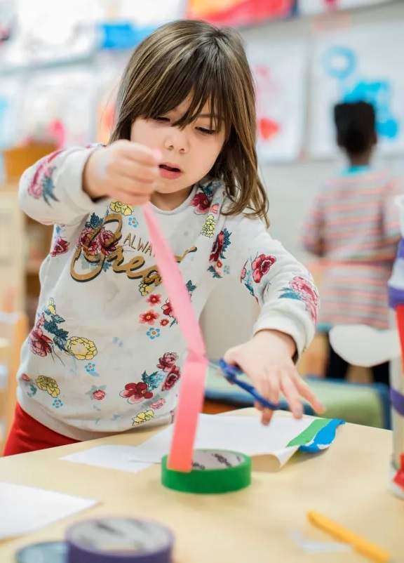 Universal prekindergarten is coming to California_bumpy rollout and all: young girl cutting tape to make art project in preschool classroom