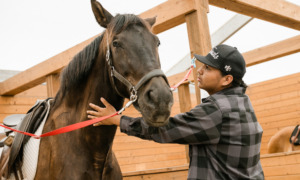 Healing Children of Horse Nations: Young man with dark hair, wearing black baseball cap and dark plaid shirt stands next to brown horse petting its neck.