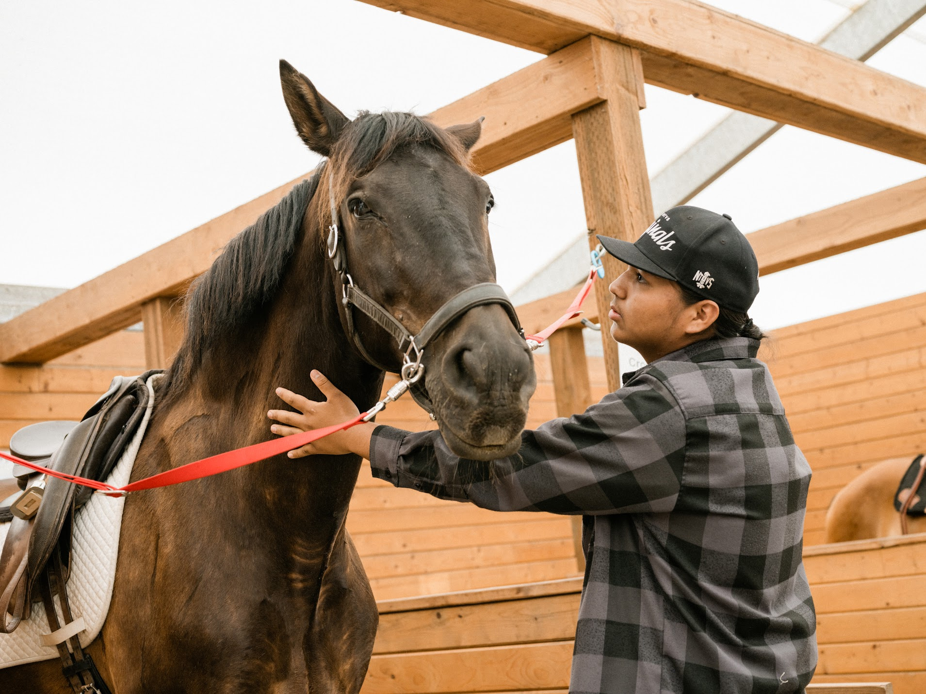Healing Children of Horse Nations: Young man with dark hair, wearing black baseball cap and dark plaid shirt stands next to brown horse petting its neck.
