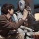 ChatGPT student cheating: Male teen with brown hair sits next to larger adult-size white humanoid robot. They are looking at each other and giving each other high five with hands meeting.