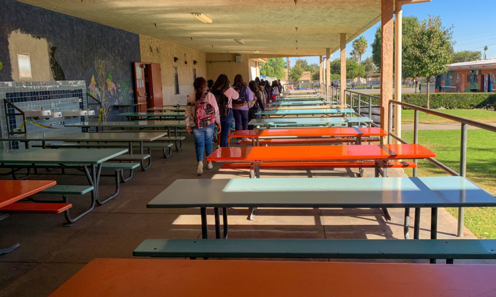 No school suspension: Two rows of many empty, long tables on pale blue or bright orange with attached benches are in a covered cemented area, with several people lined up single file in aisle between them