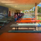 No school suspension: Two rows of many empty, long tables on pale blue or bright orange with attached benches are in a covered cemented area, with several people lined up single file in aisle between them