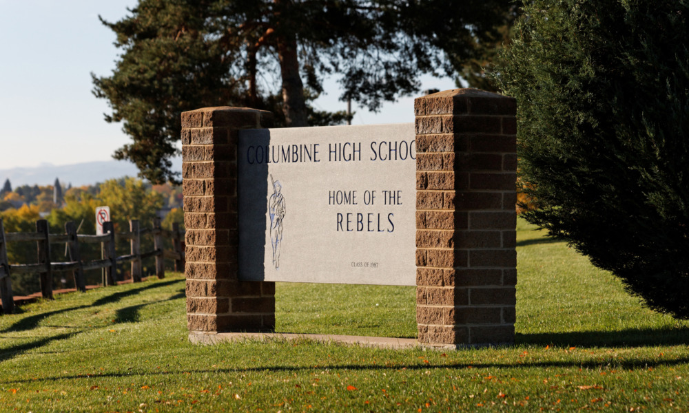 Q&A Teaching after Columbine: Columbine High School sign - cement center with red brick pillars - in 2012, on grass slope with evergreen trees in background.