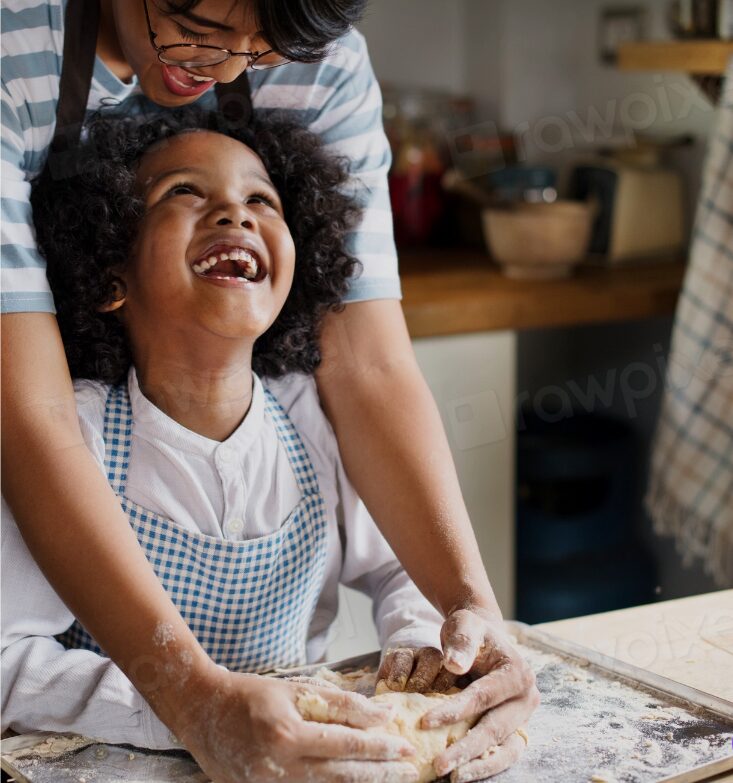 Guaranteed Income: Young black child with huge smile looks up at mother as she guides child's hands to knead bread dough covered with flour on kitchen countertop they stand next to