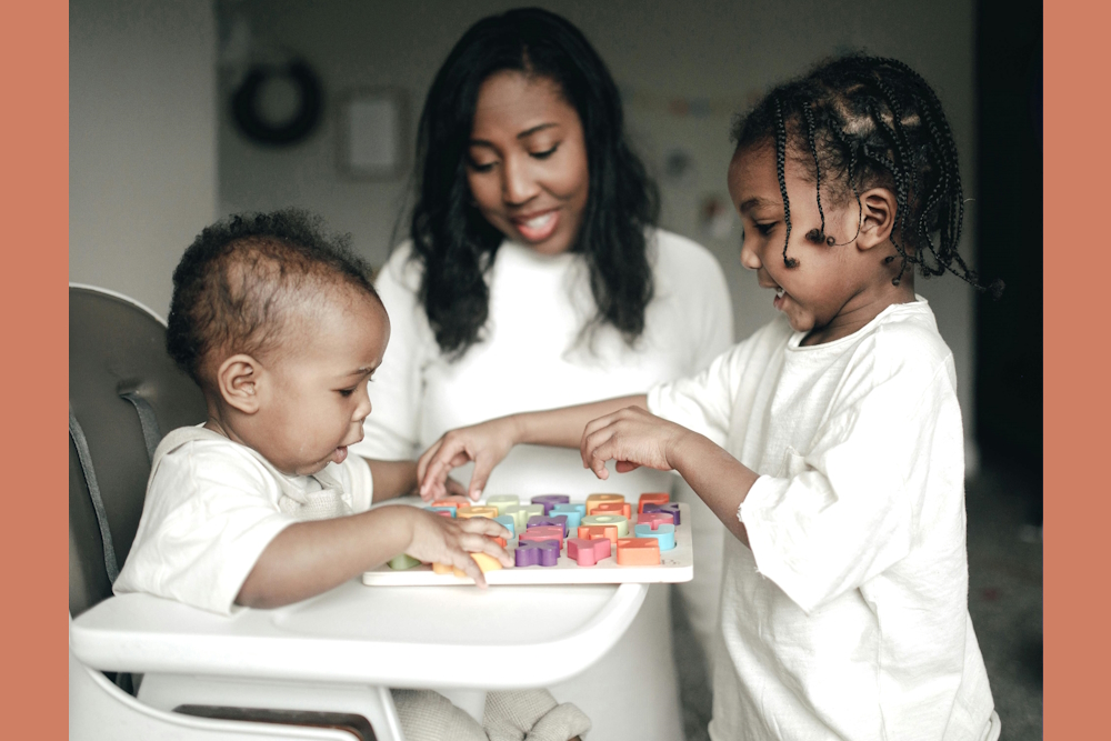 Guaranteed Income: Black mother, infant in a white high chair and toddler — all dressed in white — play with colorful 3D letter on a puzzle board on the high chair tray