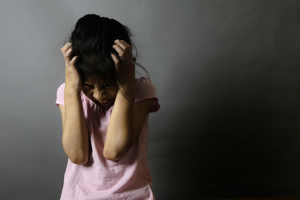 Suspension disabled students: young girl with dark hair in pink r-shirts holds head in hands with angry face against shadowed, dark background