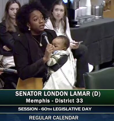 Tennessee arms teachers: Black woman with natural hairstyle in dark suit stands speaking into microphone she holds while also holding an infant wrapped in white blanket in her arms